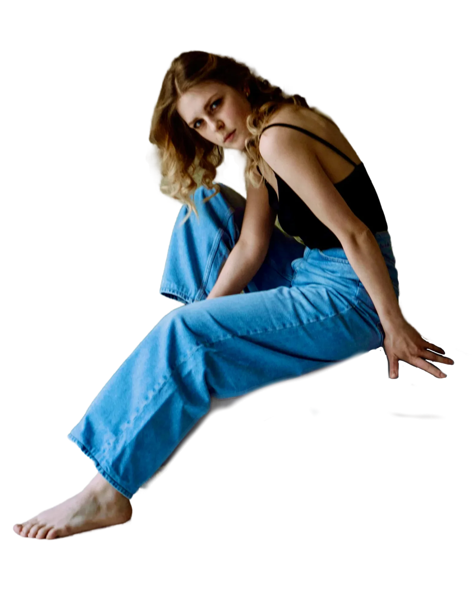 The image features a young woman in a relaxed and contemplative pose. She is seated with one leg bent and the other stretched out, barefoot, suggesting a casual and comfortable setting. Her outfit consists of a black sleeveless top with thin straps and a pair of high-waisted, loose-fitting blue jeans, giving off a chic, laid-back vibe